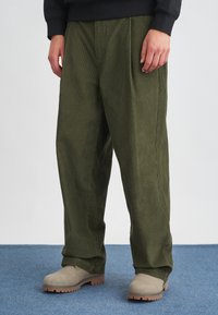 Olivno zeleni korduroj hlače s sproščenim krojem, ki imajo sprednje gube in stranske žepe, v kombinaciji s svetlo bež čevlji.