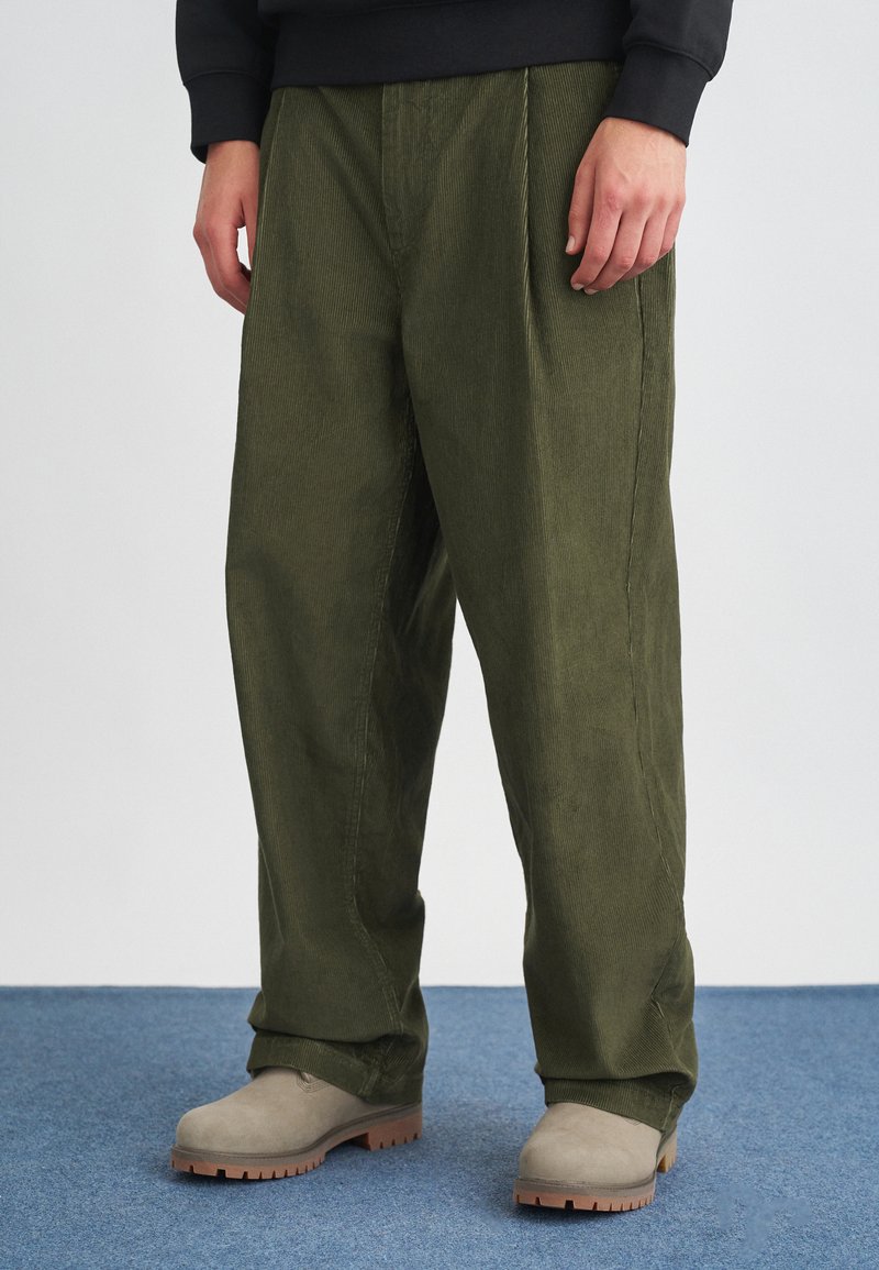 Olivno zeleni korduroj hlače s sproščenim krojem, ki imajo sprednje gube in stranske žepe, v kombinaciji s svetlo bež čevlji.