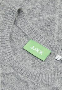 Maglione grigio lavorato a maglia con un motivo testurizzato, dotato di un'etichetta verde con "JJXX" e la taglia "S" sul colletto, materiale morbido.