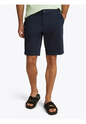 Homme portant un short bleu marine foncé avec un bouton et une fermeture éclair à l'avant, une chemise vert clair, et des sandales noires à bout ouvert, debout devant un fond blanc.