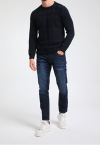 Granatowy sweter z dzianiny z wzorem diamentowym, zestawiony z ciemnoniebieskimi dżinsami i białymi trampkami. Sweter ma ściągacze na mankietach i dekolcie.