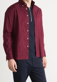 Burgundowa koszula z guzikiem i kołnierzykiem, wykonana z materiału o gładkiej fakturze. Posiada srebrny zamek błyskawiczny oraz subtelne logo na klatce piersiowej.