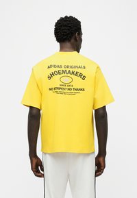 Άνδρας που στρέφεται με την πλάτη φορώντας κίτρινη μπλούζα Adidas Originals Shoemakers με μαύρο κείμενο και άσπρο παντελόνι με μαύρες ρίγες.