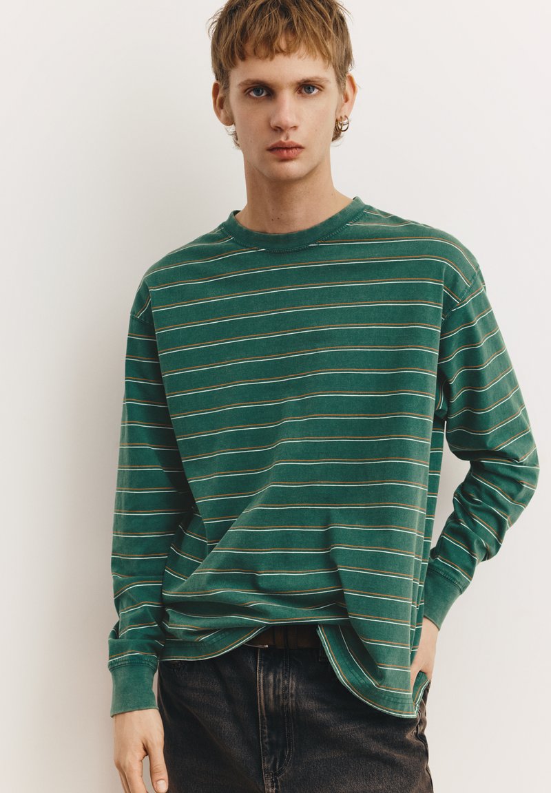 Tricou verde cu mâneci lungi, cu dungi orizontale albe și maro deschis. Guler rotund, croială lejeră, textură moale și manșete cu nervuri.