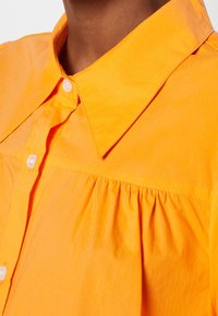 Chemise orange à col structuré, fabriquée en tissu doux, avec un design de découpe et des plis verticaux, ainsi que de petits boutons blancs.