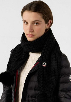 Zwarte gebreide sjaal met een gestructureerd patroon, voorzien van twee bont pompons aan de uiteinden en een ronde logo label. Een warme en veelzijdige accessoire.