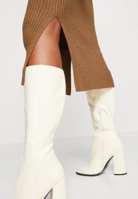Stivali bianchi fino al ginocchio realizzati in materiale liscio, con un tacco spesso e una zip laterale, abbinati a una gonna brown a costine con spacchi.