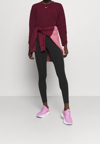 Persona che indossa un felpa bordeaux, leggings neri, scarpe da ginnastica rosa e un felpa rosa annodata in vita, su uno sfondo uniforme.