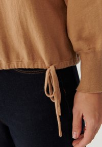 Vaaleanruskea neulottu neulepaita, jossa on teksturoitu viimeistely, sivusolmittava yksityiskohta helmassa ja resorit hihansuissa. Päällä tummansiniset farkut.