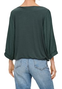 Top verde de corte holgado con un escote amplio y mangas tres cuartos. Tejido texturizado con puños enrollados. Combinado con vaqueros de color azul claro.