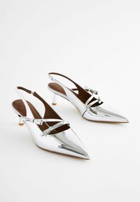 Stříbrné slingback podpatky se špičatou špičkou a lesklým povrchem, se zkříženými pásky a malým blokovým podpatkem. Hladká textura s hnědými stélkami.
