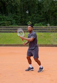 Mängija hallis spordipolis ja lühikestes pükstes hoiab tennise reketit saviväljakul. Riietust täiendavad mustad spordijalatsid ja valged sokid.