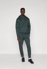 Tumši zaļa kapuce un atbilstoši sporta bikses, izgatavotas no mīksta auduma. Tās ir ar priekšējo kabatu, auklām un ribotām aprocēm. Melni sporta apavi.