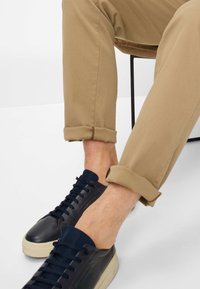 Sneaker in pelle nera con accenti blu navy, abbinati a pantaloni beige arrotolati con cuciture marroni sui polsini.