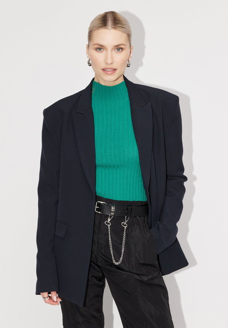 Blazer noir surdimensionné avec épaules structurées, associé à un col roulé côtelé vert. Une ceinture noire avec des accents en chaîne. Pantalon avec des plis horizontaux.