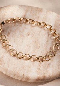 Goldene Kettenarmband mit miteinander verbundenen ovalen Gliedern und glänzender Oberfläche, auf einer glatten, hellfarbigen Steinoberfläche ruhend.