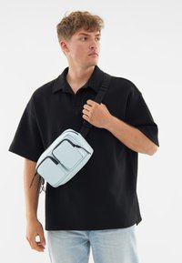 Ljusblå crossbody-väska med två framfickor, justerbar svart rem och strukturerad yta. Bärs av en person med svart polo-tröja.