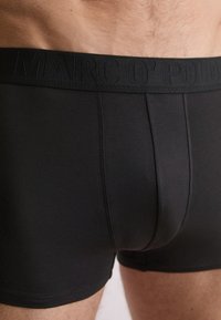 Primo piano di una persona che indossa boxer neri con una cintura elastica nera impreziosita da un sottile lettering del marchio.