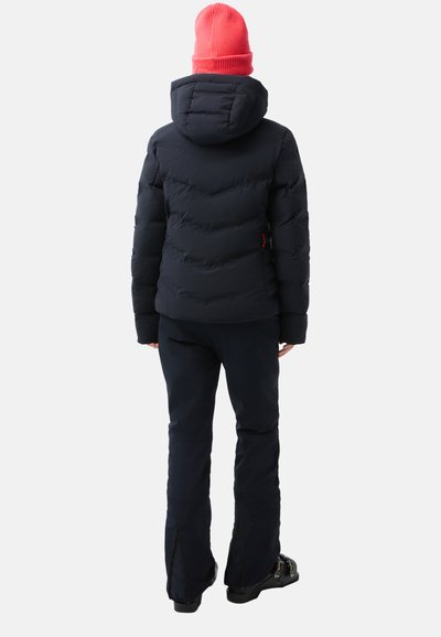 Navyblauer Pufferjacke mit Kapuze, strukturiertem Steppmuster; kombiniert mit schwarzen Hosen mit Reißverschlüssen an den Knöcheln und schwarzen Skistiefeln; leuchtend rosa Mütze.