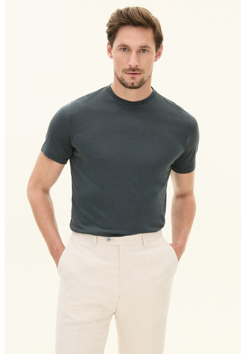 Man met lichtbruin haar, gekleed in een donkergrijs kortemouwen shirt en lichtbeige broek, staat met handen in zijn zakken tegen een effen achtergrond.