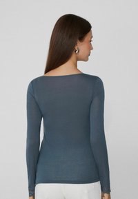 Dunkelbluse mit langen Ärmeln, die einen Bootsausschnitt und transparente Spitze an den Bündchen hat. Glatte Textur mit taillierter Silhouette.