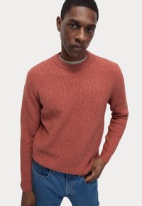 Suéter de punto rojo con un patrón texturizado, escote redondo y puños de canalé, combinado con jeans azules. Fondo liso.