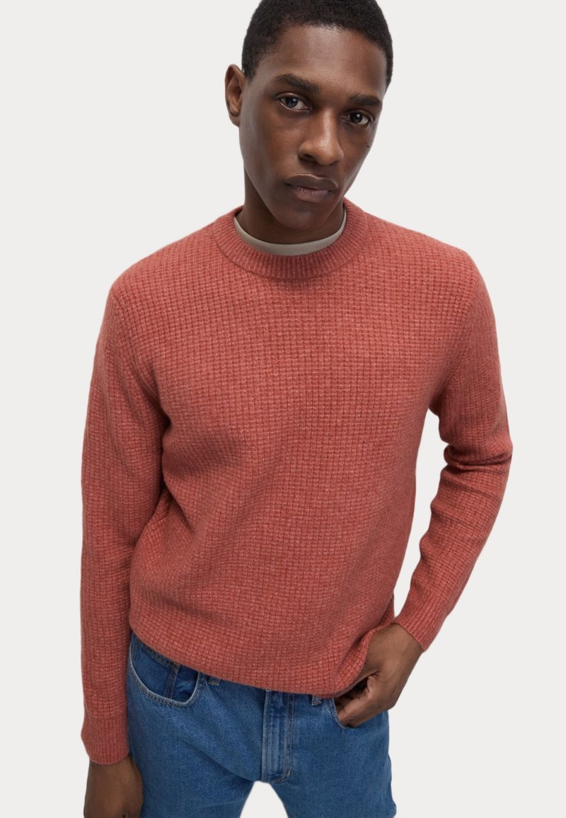 Suéter de punto rojo con un patrón texturizado, escote redondo y puños de canalé, combinado con jeans azules. Fondo liso.