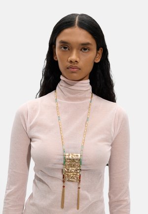 Młoda kobieta z długimi ciemnymi włosami, ubrana w przezroczysty blado-różowy golf i długi naszyjnik z złotych, zielonych i czerwonych koralików z przywieszką chwostu.