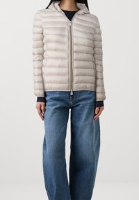 Helle beigefarbene Daunenjacke mit Reißverschluss vorne, Kapuze und gestepptem Design. Lange Ärmel und eine figurbetonte Silhouette, kombiniert mit weit geschnittenen blauen Jeans.