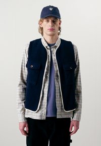 Tumši zils veste ar četriem priekšējiem kabatām, gaiši brūna apdare, uzlikta virs rakstainā krekla. Modelis valkā tumšu cepuri un violetu T-kreklu zemāk.