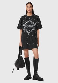 Donna che indossa un abito t-shirt oversize nero con stampa a catena bianca, tiene una borsa nera e indossa stivali neri fino al ginocchio.
