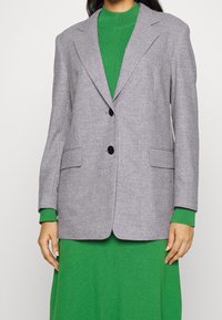 Blazer gris à deux boutons, avec des revers crantés et deux poches avant. Assorti à un col roulé côtelé vert et une robe en tricot. Texture lisse.
