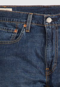 Jeans de denim azul oscuro con bolsillos delanteros, remaches de cobre, costuras naranjas, trabillas para el cinturón y un botón metálico en la cinturilla.