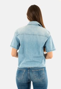 Camisa de mezclilla de manga corta en azul claro con cuello, que presenta una costura trasera recta y una textura ligeramente desgastada. Combinada con jeans azules.