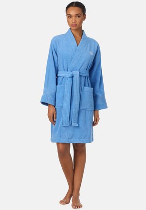 Vrouw staat blootsvoets met een blauwe badstof badjas tot op de knie met zakken en een taillekoord tegen een witte achtergrond.