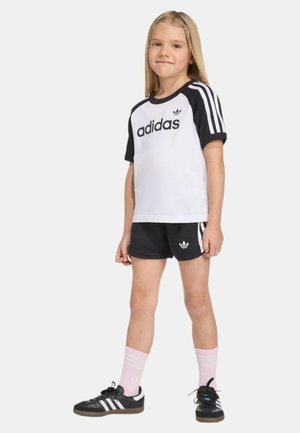 Ragazza giovane in piedi, indossa abbigliamento sportivo Adidas bianco e nero, pantaloncini neri, calzini rosa e sneakers Adidas nere con strisce bianche.