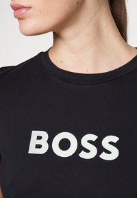 Camiseta de algodón negra con cuello redondo y un prominente logo blanco "BOSS" en el centro. Textura suave con mangas cortas.