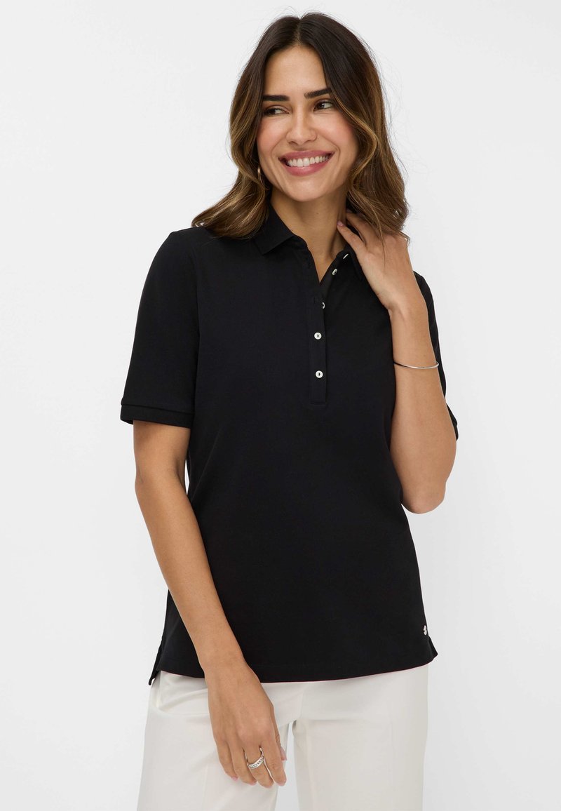 Sort polo-shirt med korte ærmer og knaplukning. Blødt stof, sideslids og diskret branding i kanten. Bæres med lette bukser.