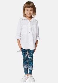 Fehér, felgöngyölt ujjú ing, kék mintás leggings-szel párosítva, amelyeken madarak és növényzet látható. A szettet fehér sportcipő zárja le.