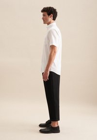 Witte short-sleeve shirt met een button-down kraag, gecombineerd met zwarte cropped broek en zwarte schoenen. Model wordt zijaanzicht getoond.