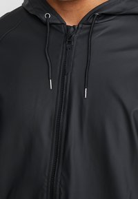 Czarna wodoodporna kurtka z zamkiem błyskawicznym z przodu, regulowanym kapturem i troczkami. Gładkie, matowe wykończenie z minimalnymi detalami szycia.