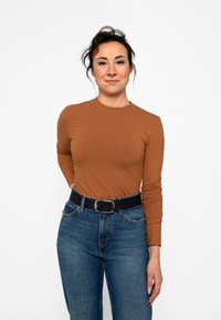 Giovane donna con capelli scuri che indossa un top marrone a costine a maniche lunghe e jeans blu con cintura nera, sorridente e in piedi davanti a uno sfondo bianco.