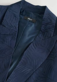 Navy blue blazer with a textured palm leaf pattern, featuring a smooth satin lining and a tag labeled "NEXT" on the inside collar.