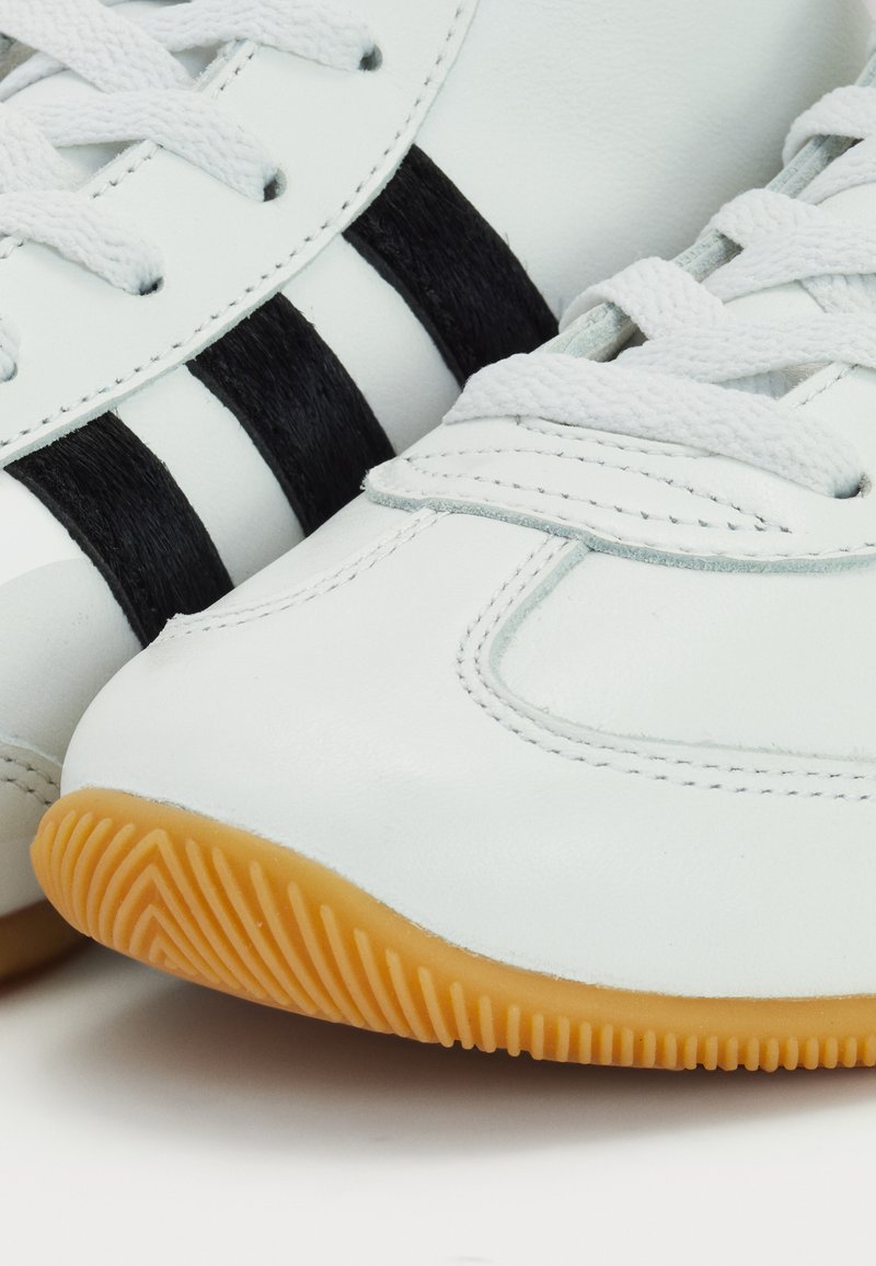 Weiße Ledersneaker mit drei schwarzen Streifen, strukturiertem Gummisohle und traditioneller Schnürung. Abgerundete Spitze mit Nahtdetails.