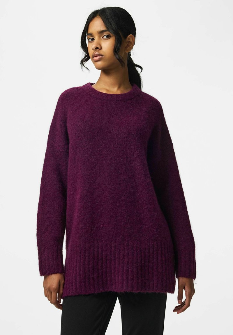 Jeune femme aux cheveux foncés attachés portant un pull violet foncé texturé et surdimensionné ainsi qu’un pantalon noir sur un fond uni.