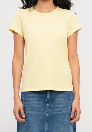 Kvinde iført en ensfarvet lysegul kortærmet T-shirt og en blå denimnederdel, stående med arme afslappet ved siden.