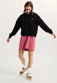 Czarna bluza z kapturem o luźnym kroju, zestawiona z różową spódnicą do kolan. Brązowe adidasy z białymi akcentami oraz widoczne białe skarpetki.