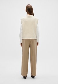 Colete de malha bege com gola alta, mangas curtas, barra canelada, usado sobre uma camisa branca de manga longa e combinado com calças largas em tom bege.