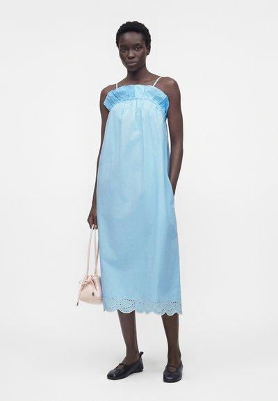 Ψηλή γυναίκα με αμάνικο μεσομήκες φόρεμα γαλάζιου χρώματος με βολάν στο ντεκολτέ, κρατώντας μικρή απαλό ροζ τσάντα, φορώντας μαύρα φλατ παπούτσια, στέκεται μπροστά σε λευκό φόντο.