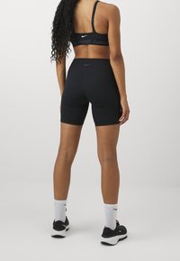 Kobieta w czarnym sportowym biustonoszu Nike, czarnych szortach rowerowych, białych skarpetkach crew Nike i czarnych sneakersach Nike, stojąca plecami na jednolitym tle.